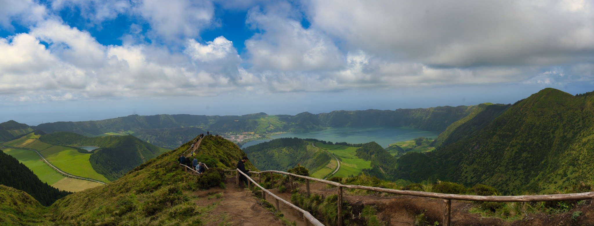 A mountain path overlooking a lake, green volcanic ridges and a wide, cloud-filled sky.