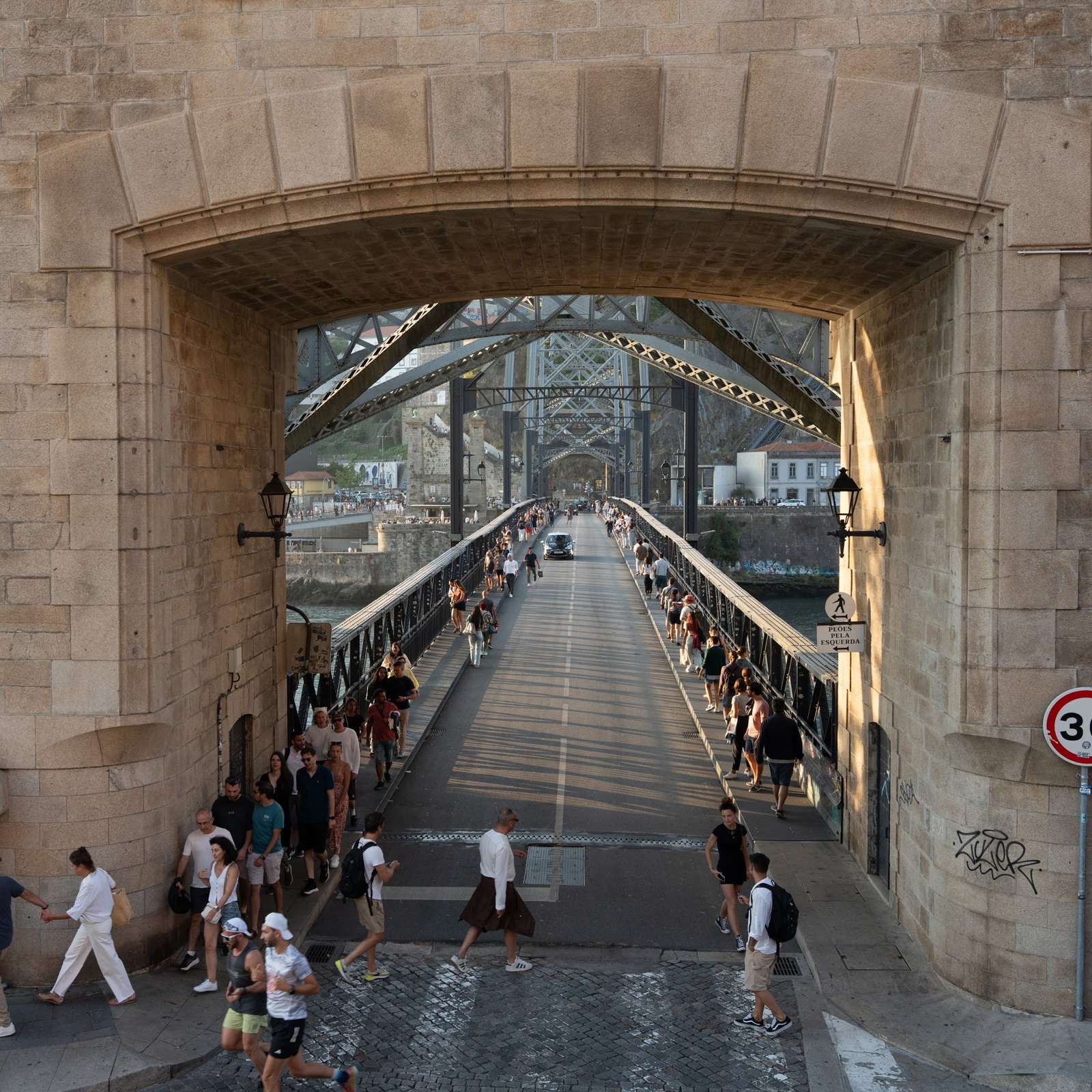 Pedestrians cross the lower deck beneath a stone arch and repeating metal beams.