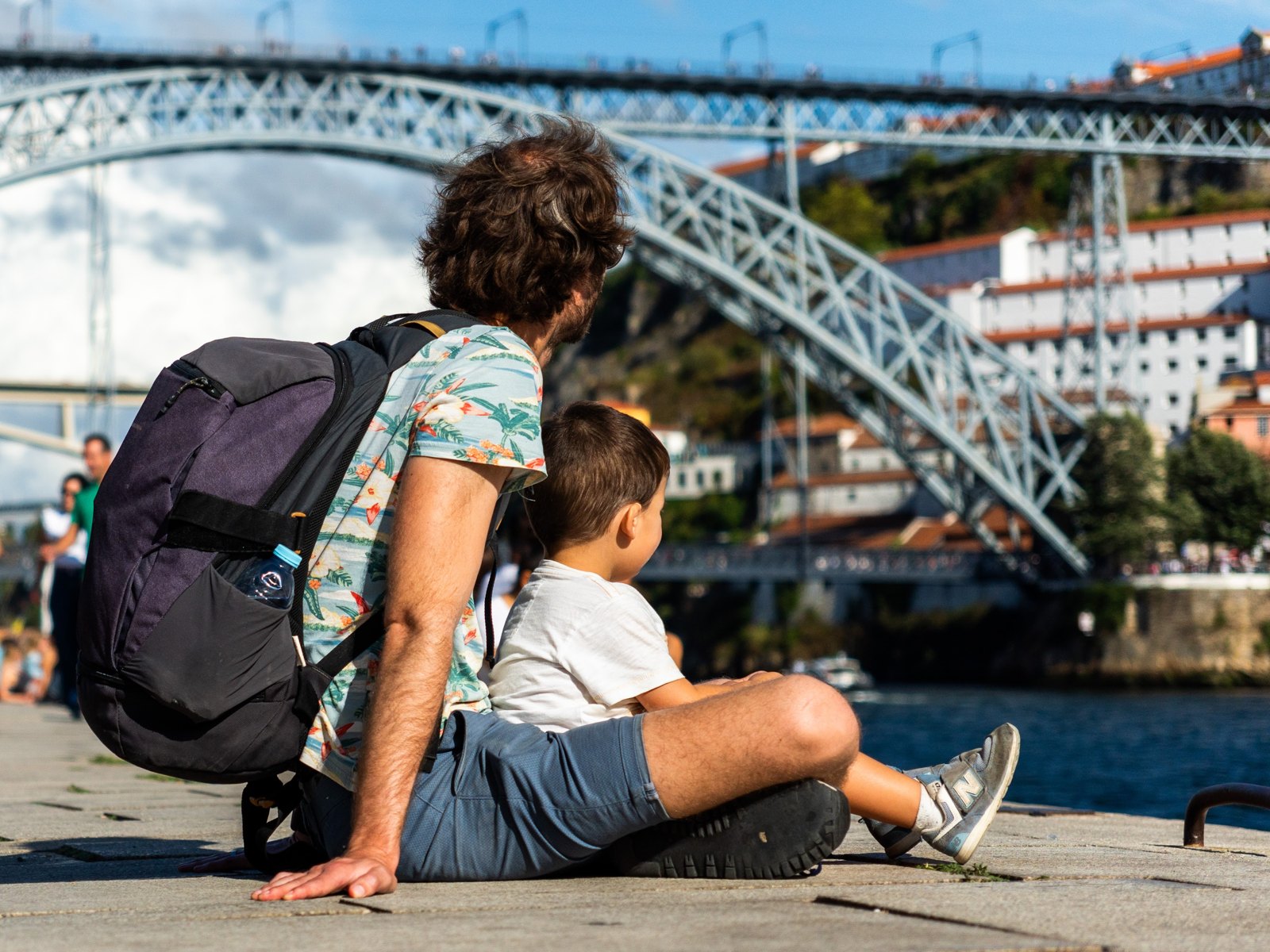 An adult and a child sit by the river looking towards the bridge.